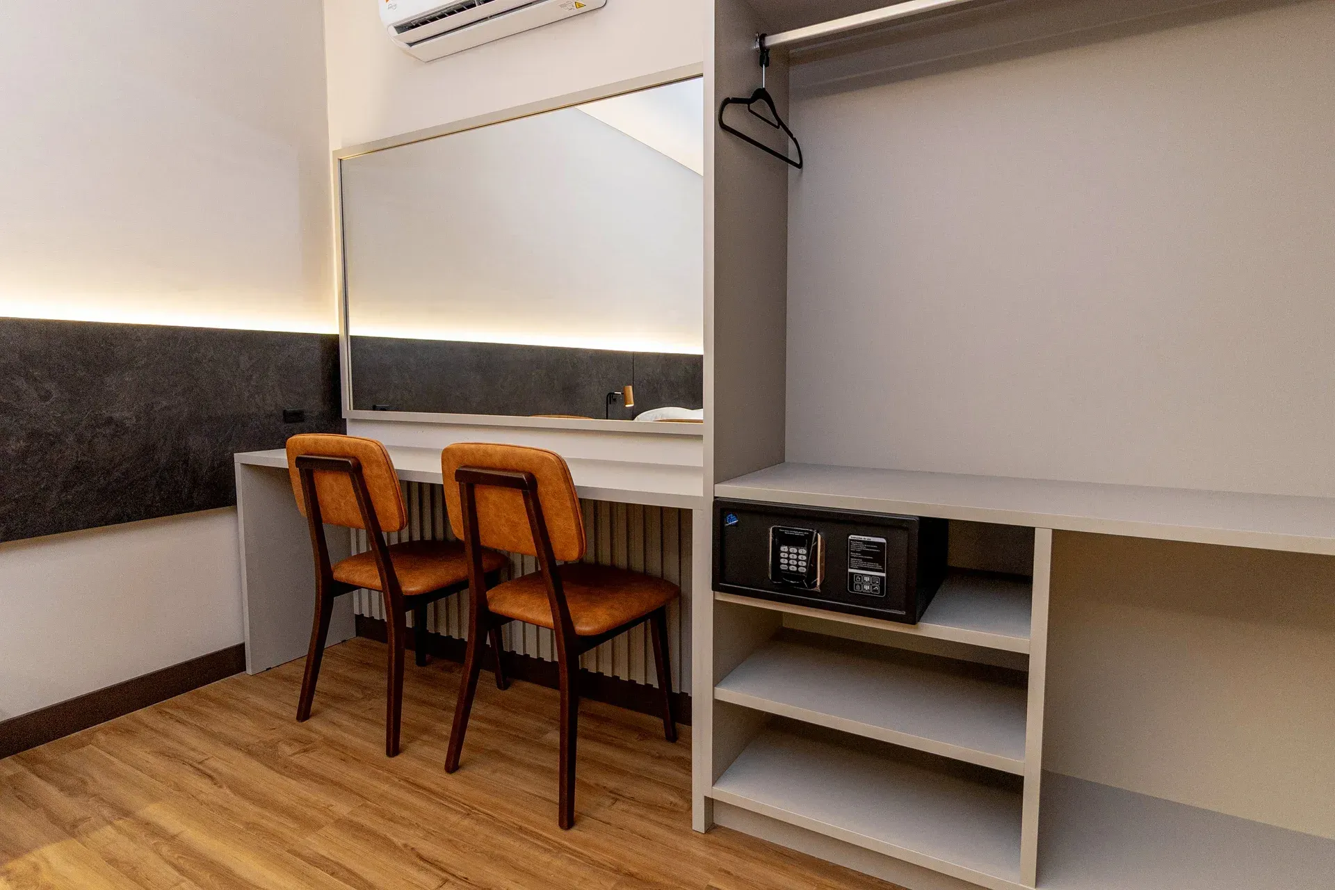 Um quarto de hotel com duas cadeiras, uma escrivaninha, um espelho e um cofre.