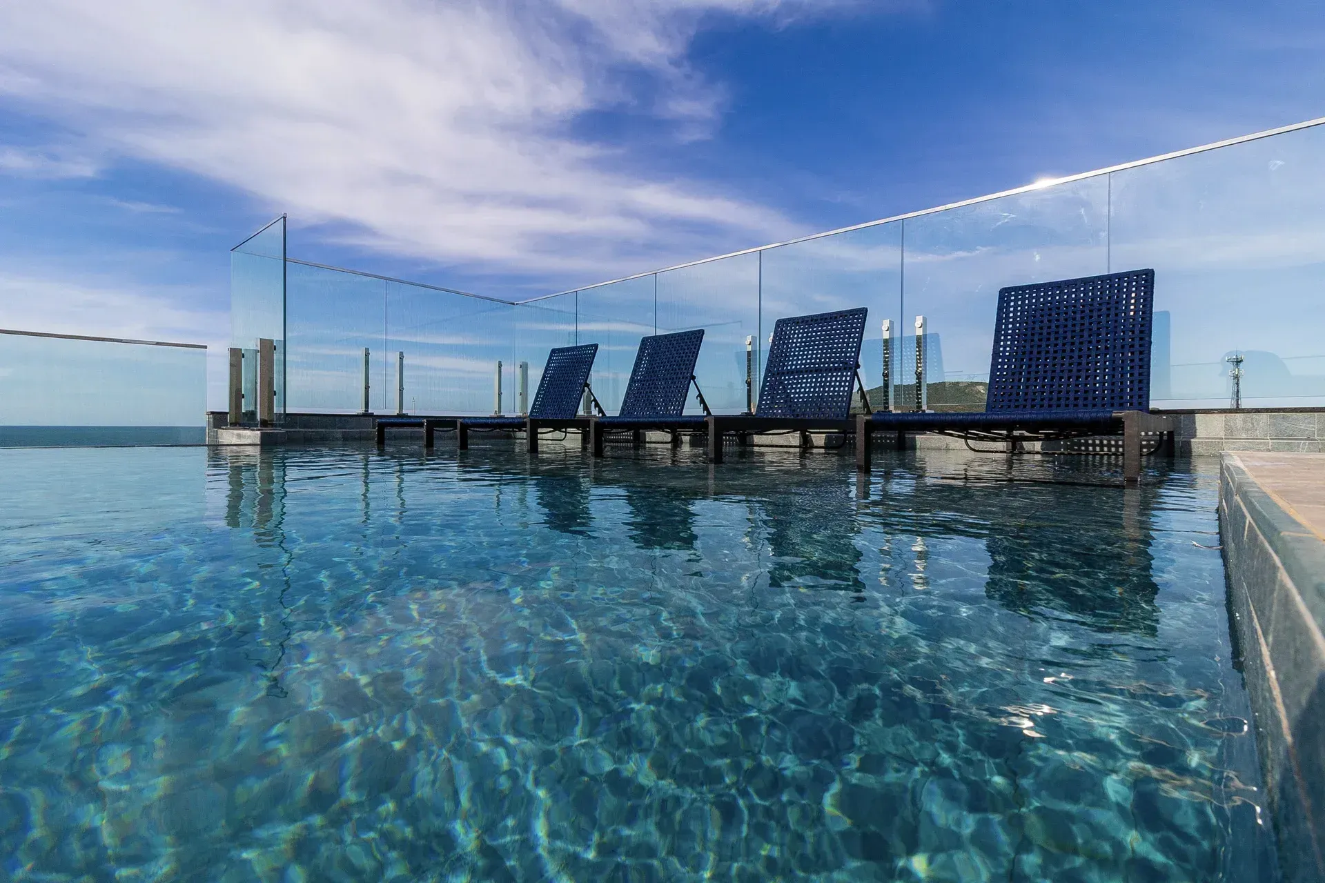 Uma piscina cercada por cadeiras e uma cerca de vidro com vista para o oceano.