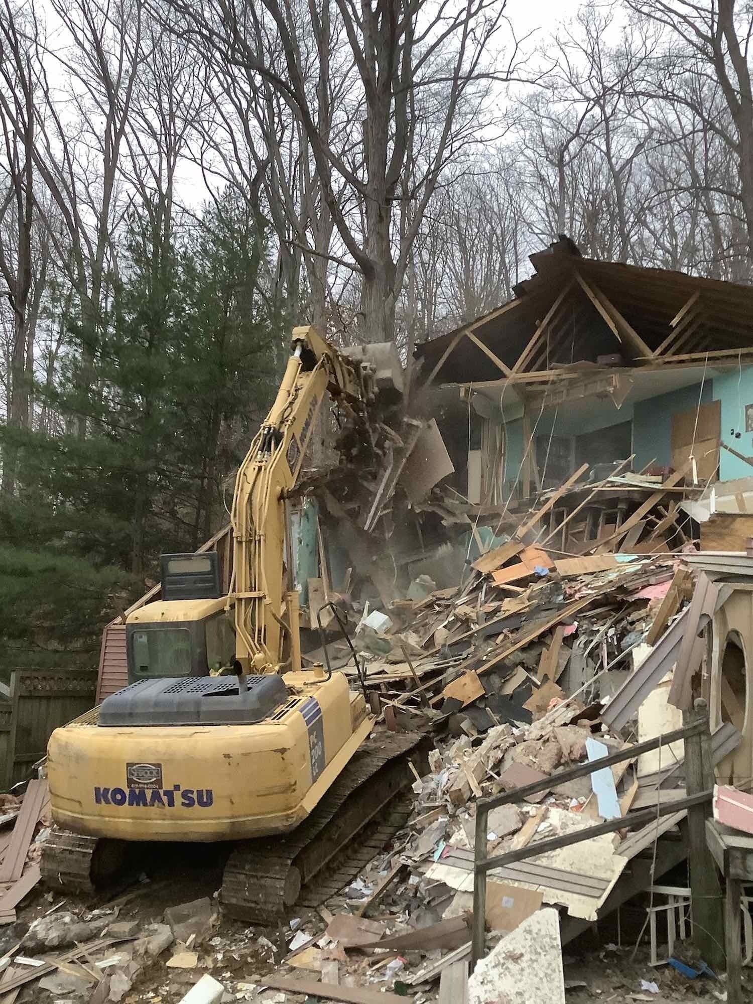 A yellow excavator is demolishing a house in the woods.