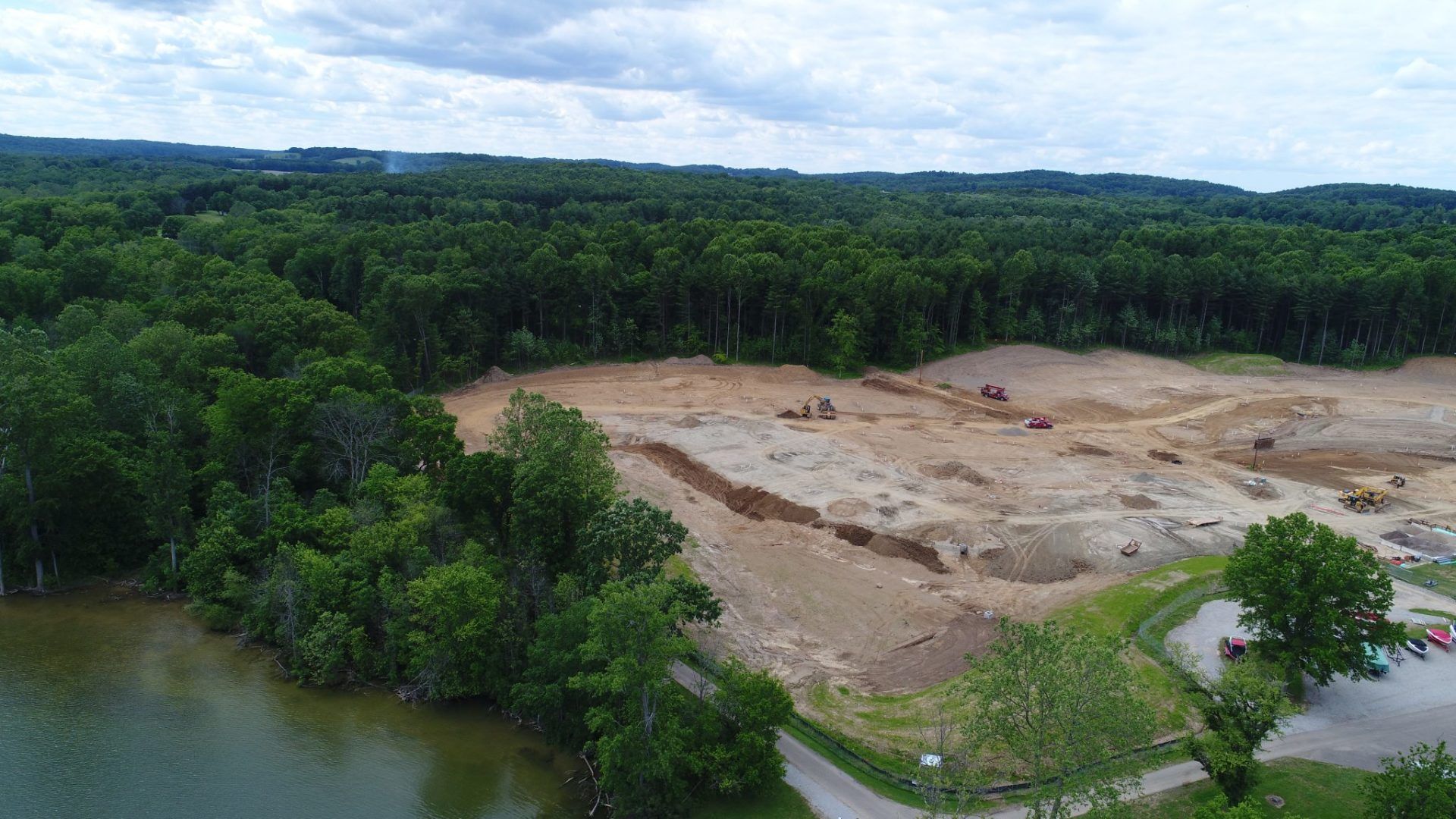 An aerial view of a dirt field next to a lake surrounded by trees.