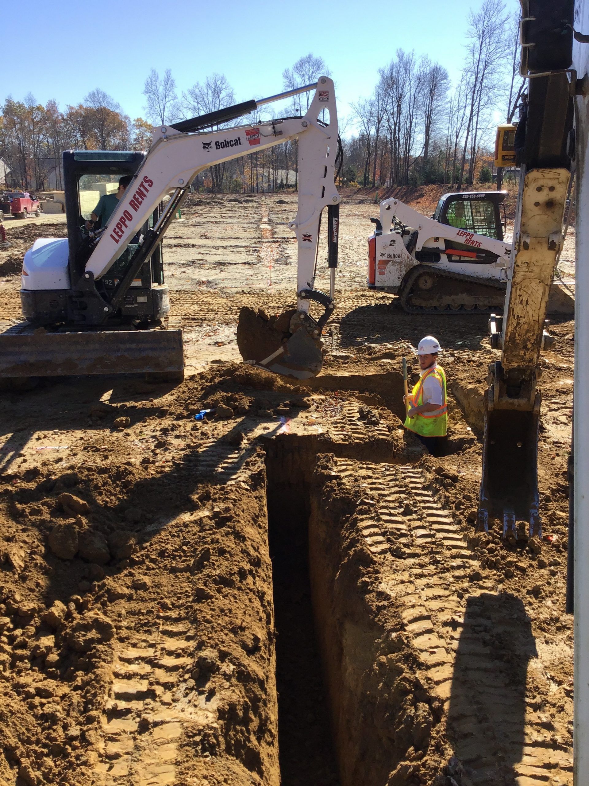 A man in a yellow vest is kneeling in the dirt next to a bobcat excavator.