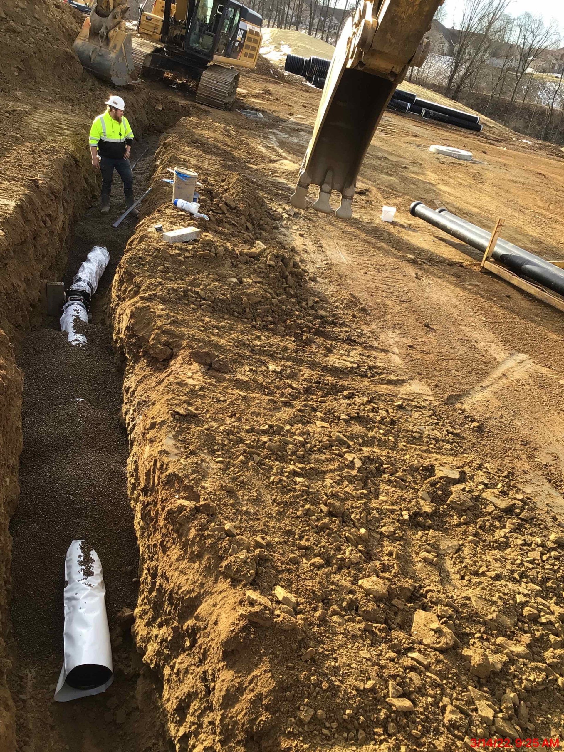 A man is standing in a trench next to a pipe in the dirt.