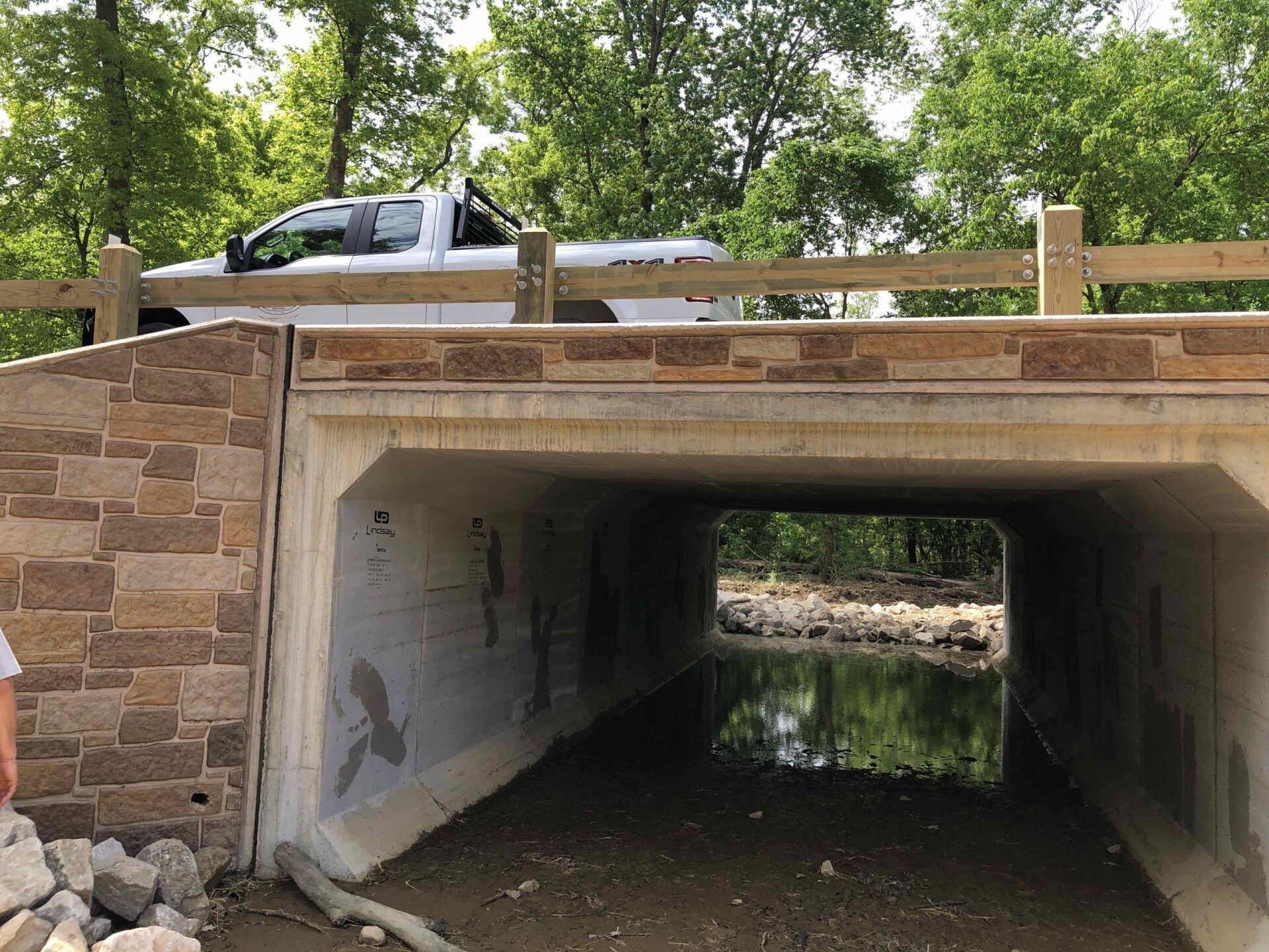 A truck is driving under a bridge over a river.