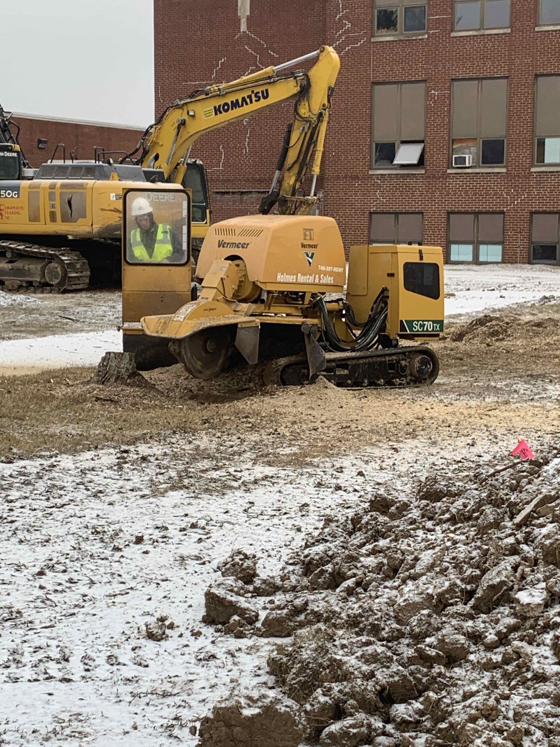 A yellow excavator is digging a hole in the dirt in front of a brick building.