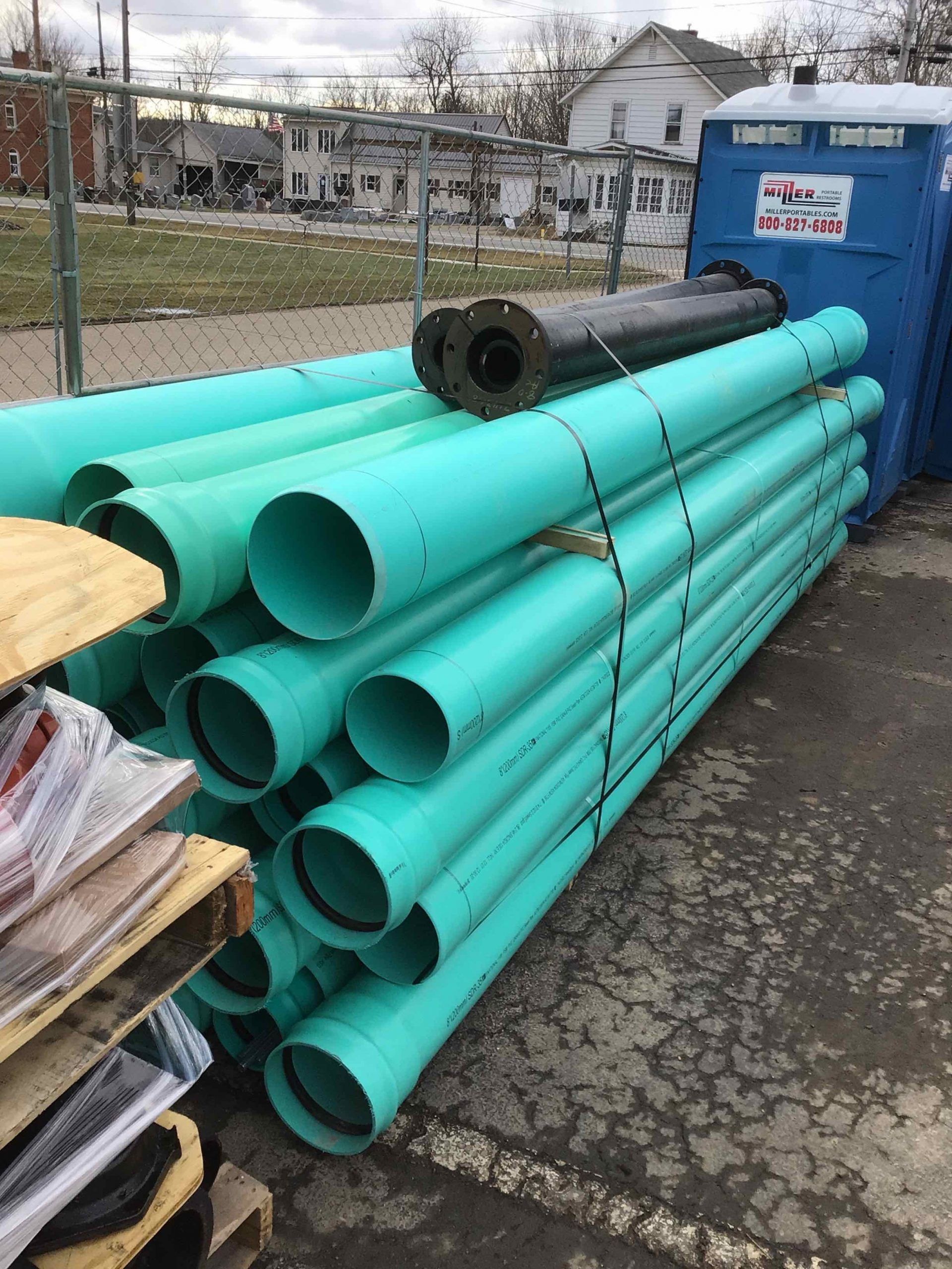 A pile of green pipes is sitting next to a blue portable toilet.