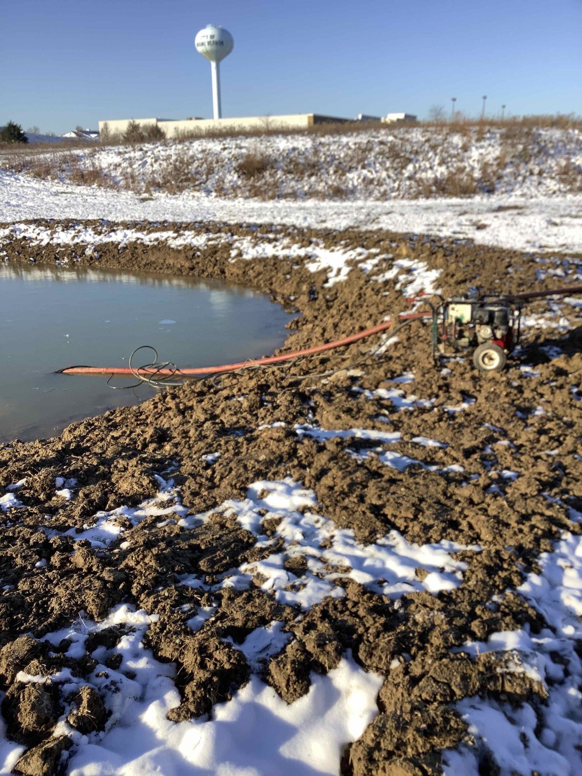 A small pond in the middle of a snowy field with a water tower in the background.