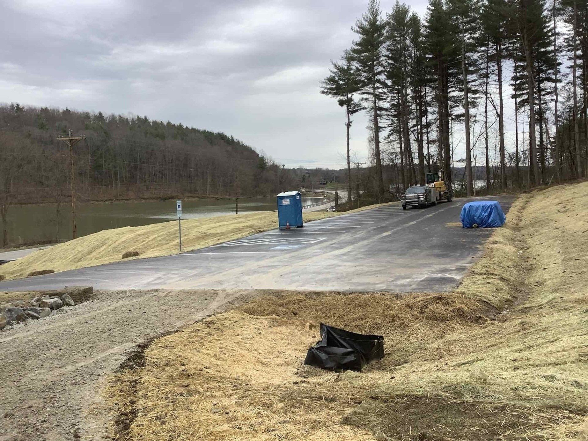 A blue portable toilet is sitting on the side of a road next to a lake.