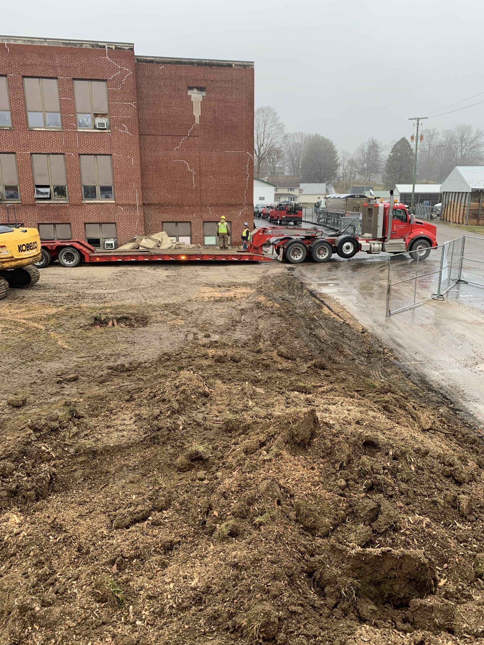 A red truck is driving down a dirt road in front of a brick building.