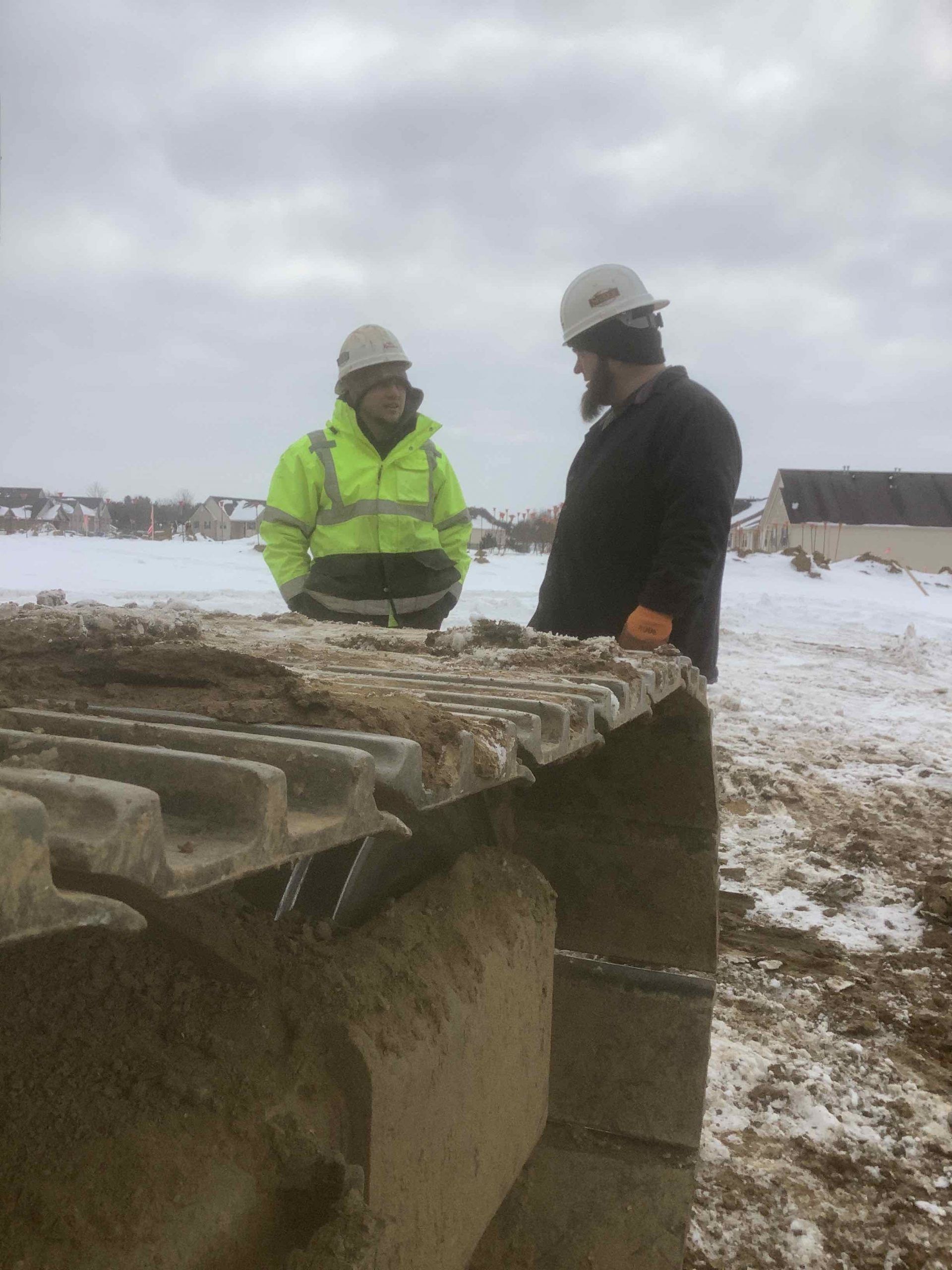 Two construction workers are standing next to a large excavator in the snow.