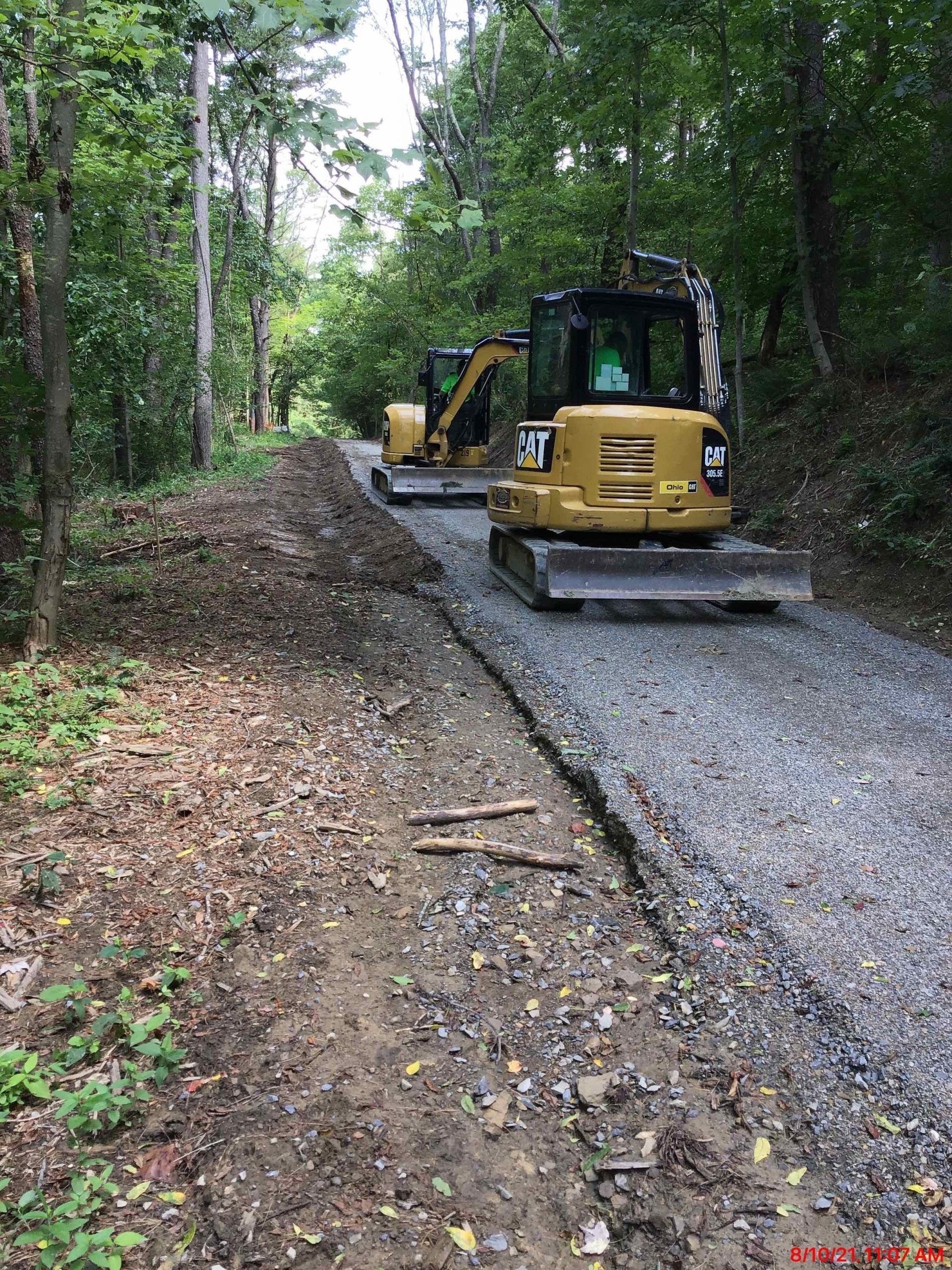 Two bulldozers are driving down a dirt road in the woods.