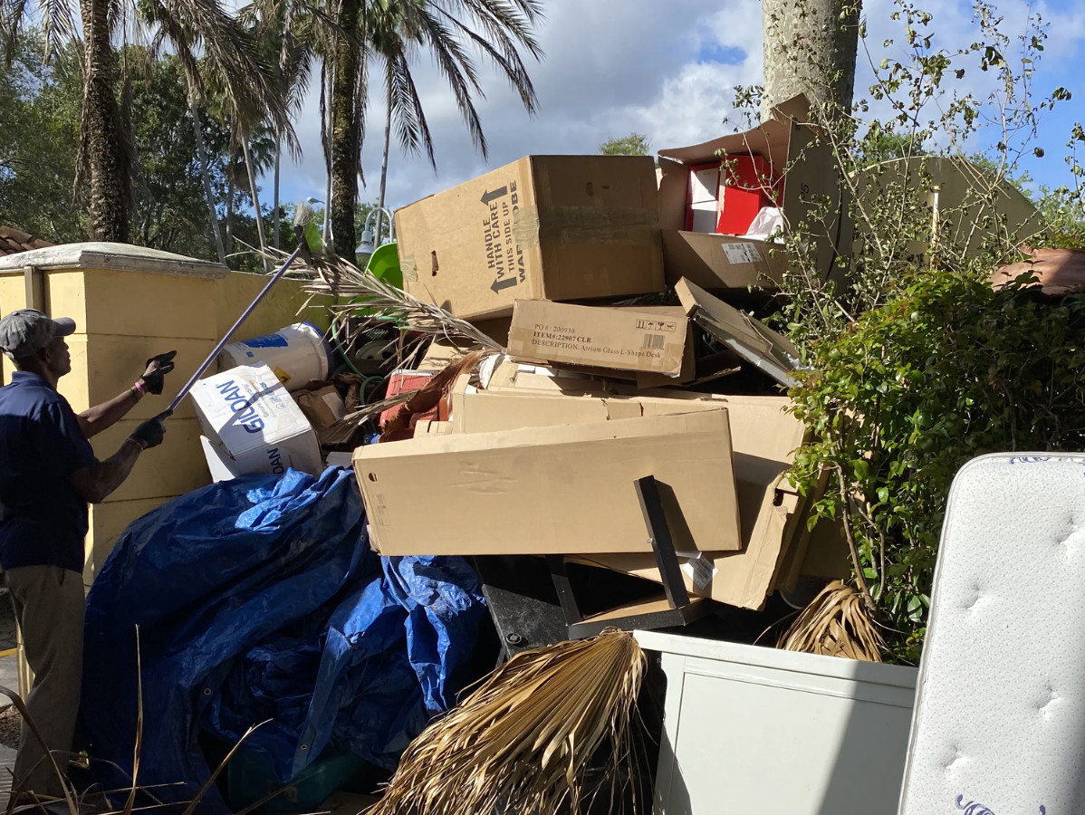 Person surveys a large pile of debris, including boxes and a mattress, next to a concrete wall.