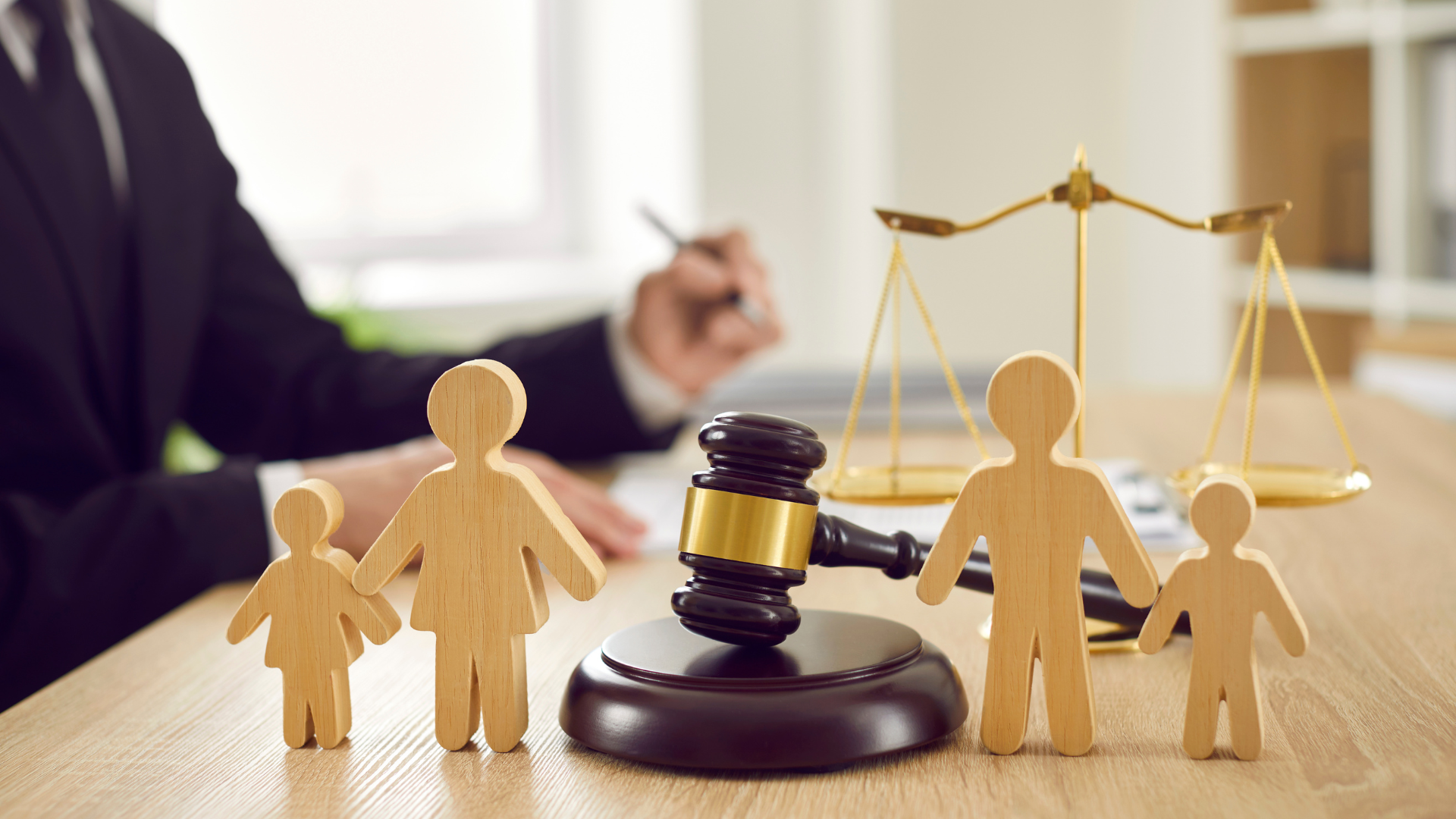 Wooden figures and a gavel beside scales of justice on a desk, with a blurred lawyer in the background