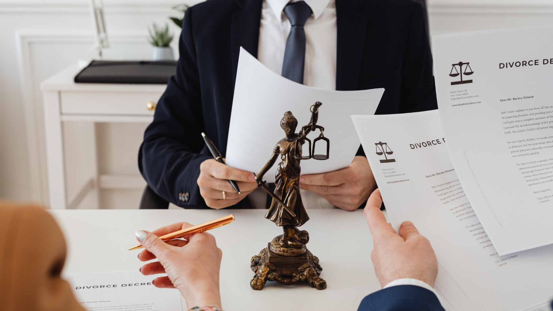 Desk meeting with lawyer reviewing papers and a bronze Lady Justice statue over documents with a pen and hand in foreground