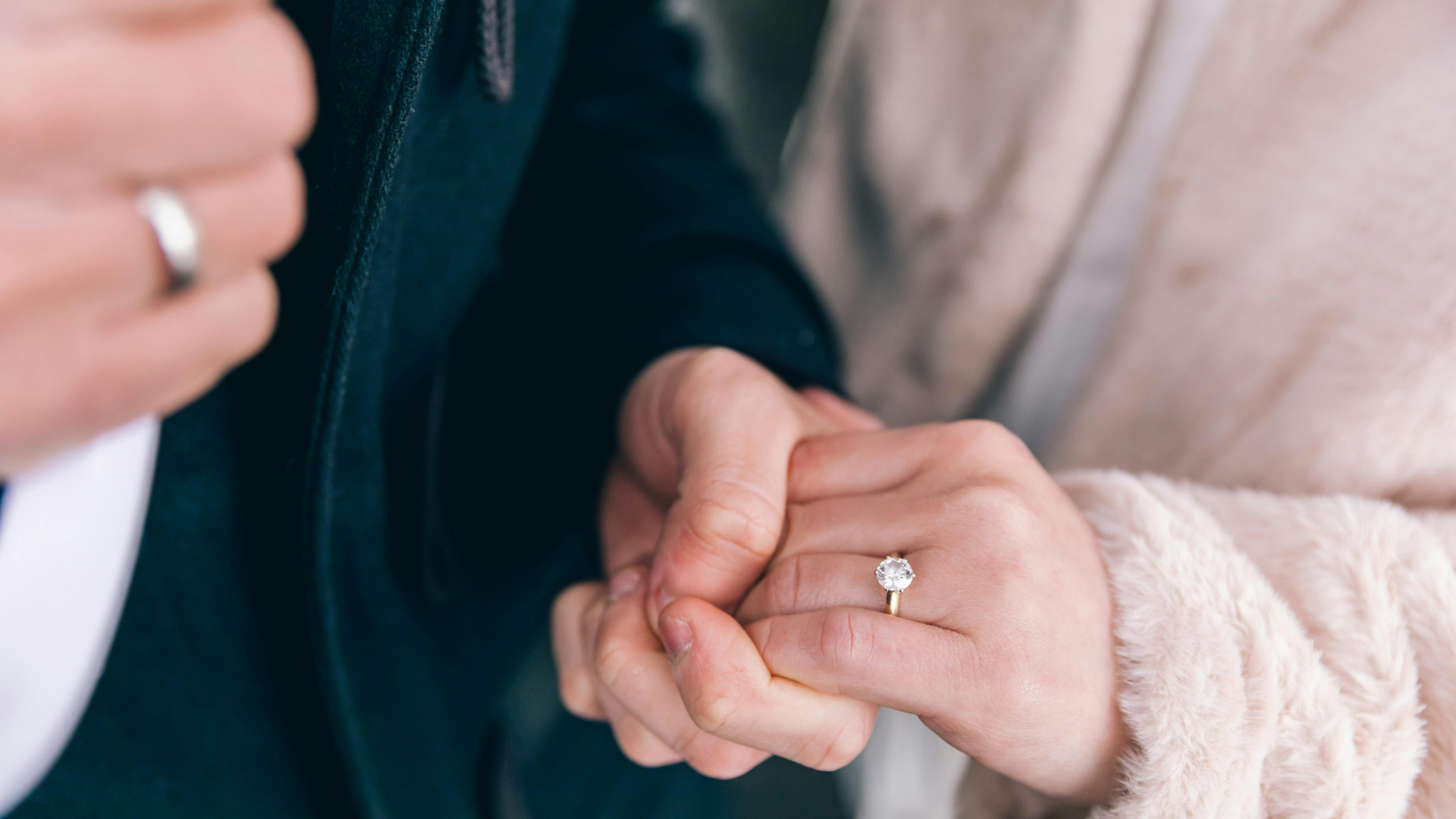 Two people holding hands, one wearing a diamond engagement ring and soft pink sweater