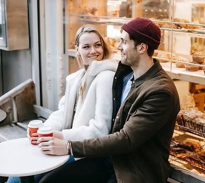 Two people sit at a café table holding coffee cups, smiling beside a bakery display.