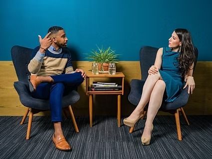 Two people sit in armchairs facing each other with a small table between them in a modern lounge.