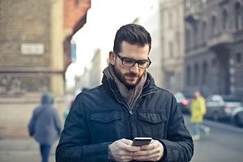 Man in a dark jacket and glasses looking at a smartphone on a city street