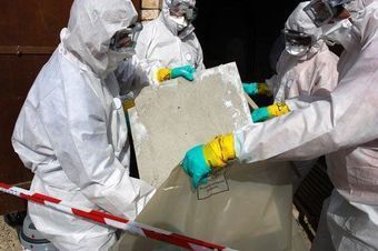 Workers in white hazmat suits handling a large concrete slab behind caution tape