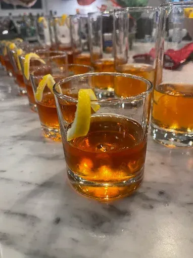 Cocktails on a marble counter, garnished with lemon twists. Brown liquid in glass tumblers, bar setting.