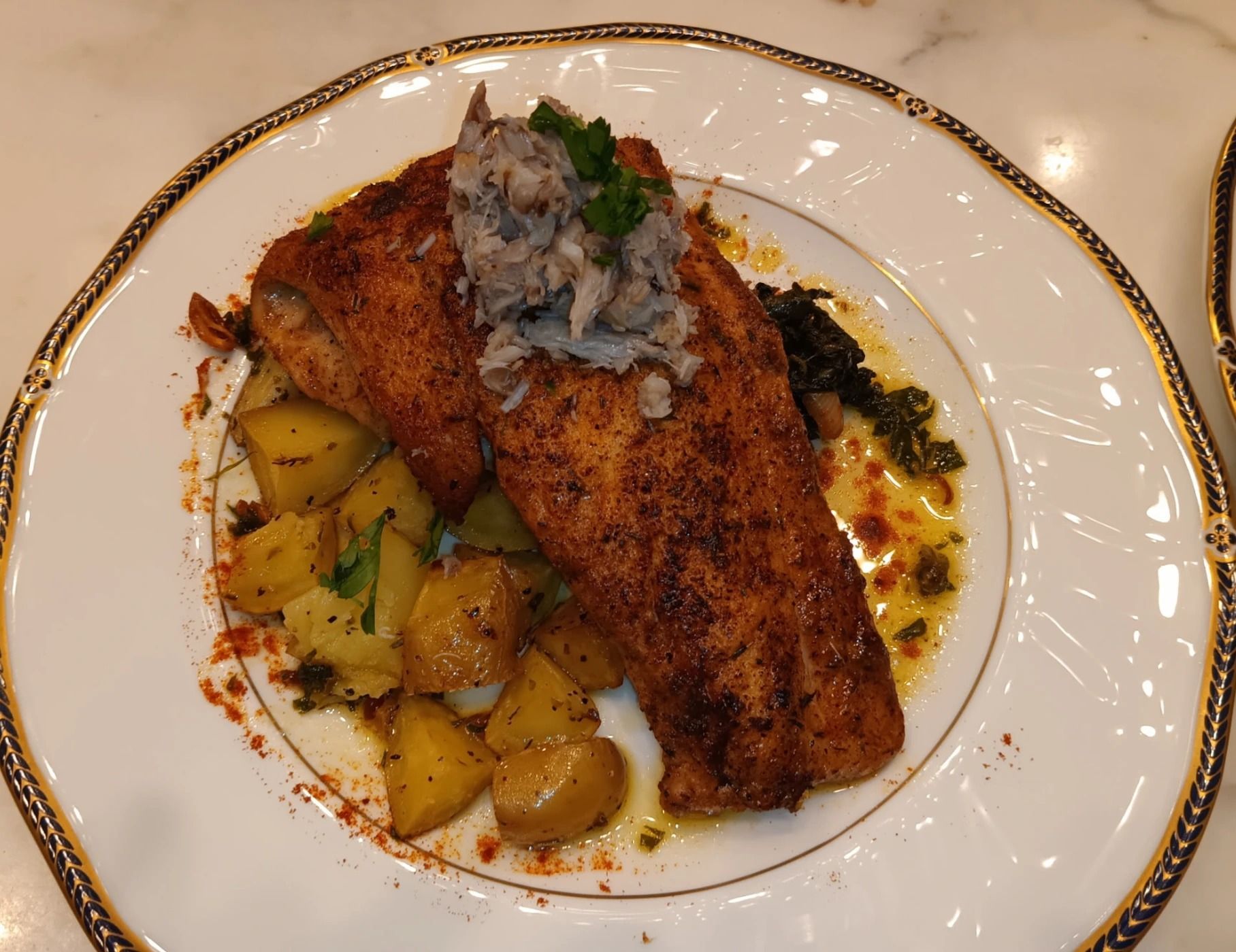 Pan-seared blackened fish fillet with potatoes and a creamy topping on a decorative white plate.