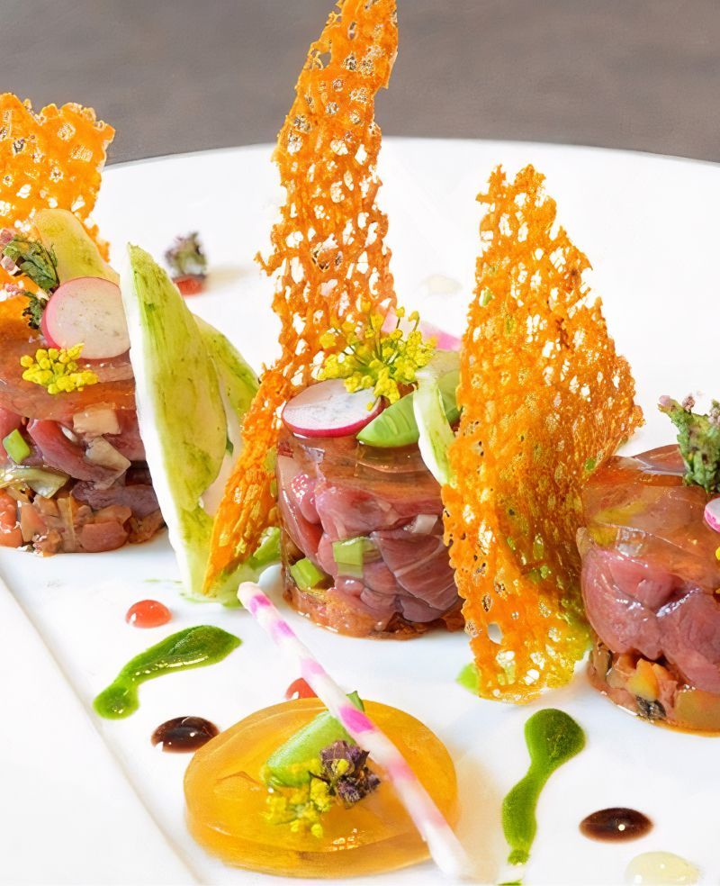 Gourmet plate with three tuna tartare stacks, orange crisps, colorful garnishes, and sauces.