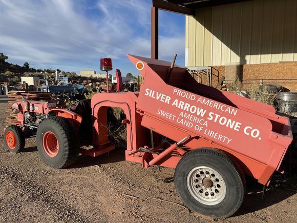 Red tractor pulling a red Silver Arrow Stone Co. trailer in a gravel lot under a partly cloudy sky