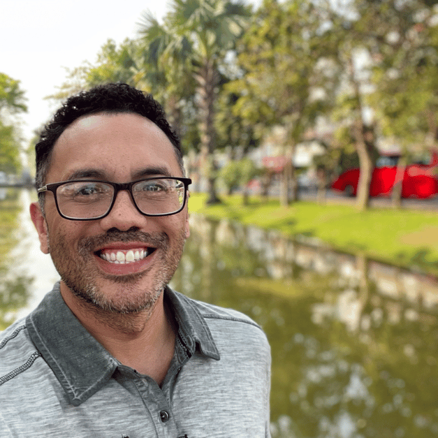 A man wearing glasses is smiling in front of a body of water.