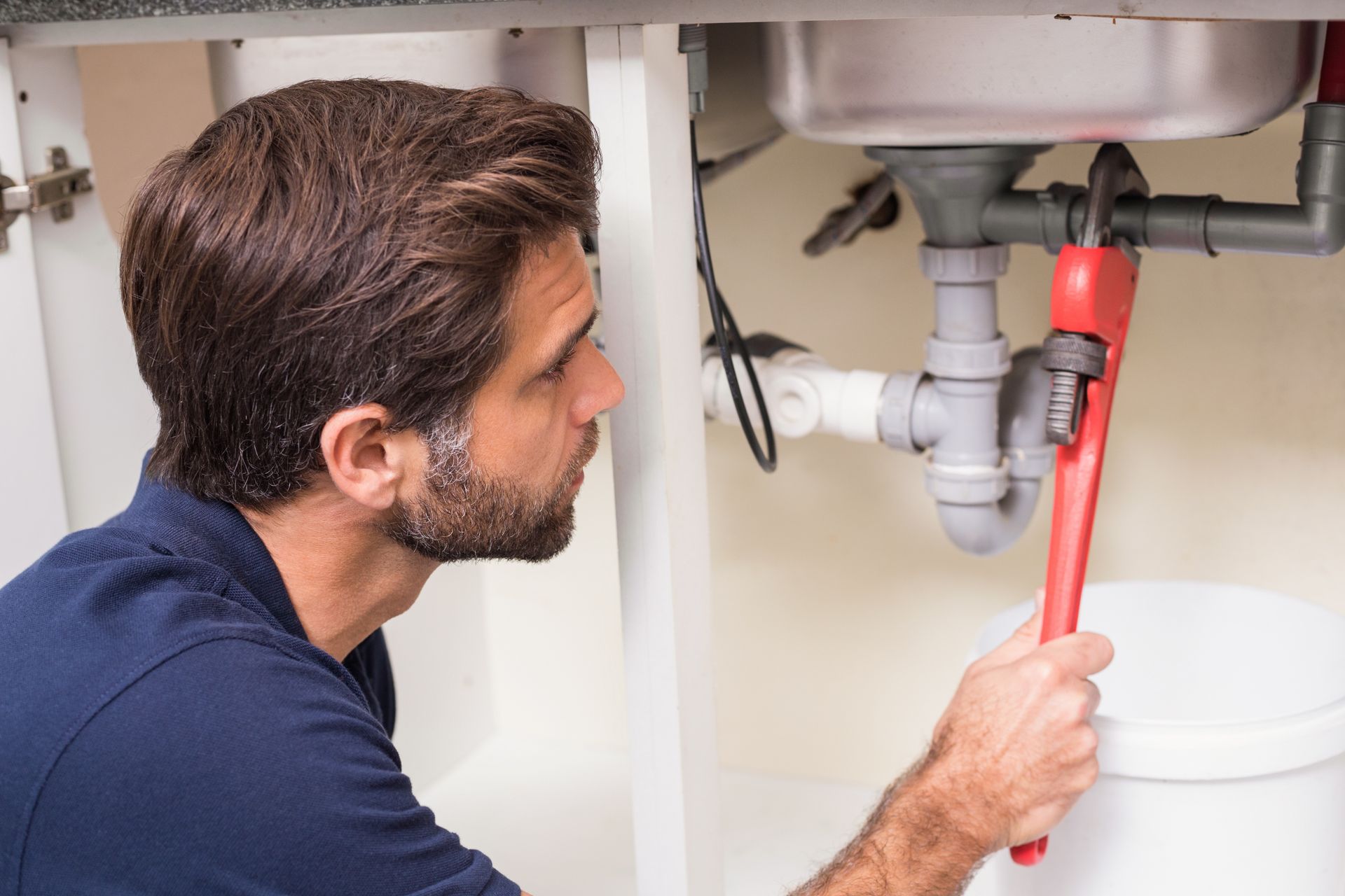 A plumber kneels under a sink, repairing plumbing with tools.