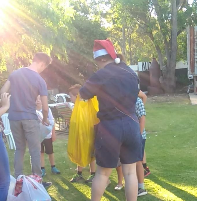 People in a yard. One wears a Santa hat and holds a yellow bag. Others gather around.