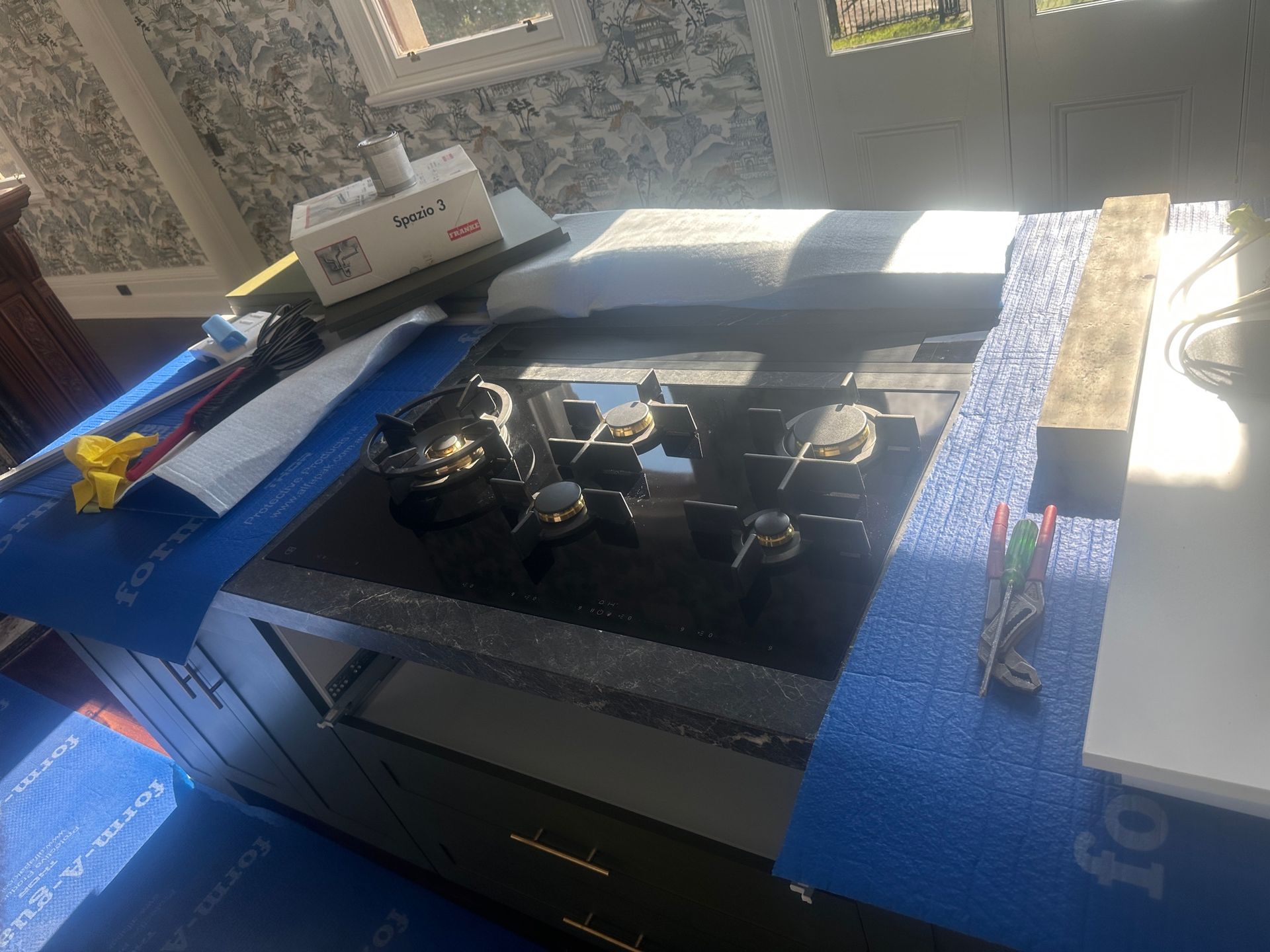 Black stovetop on a cabinet, covered with blue protective sheeting; tools and equipment on the counter.