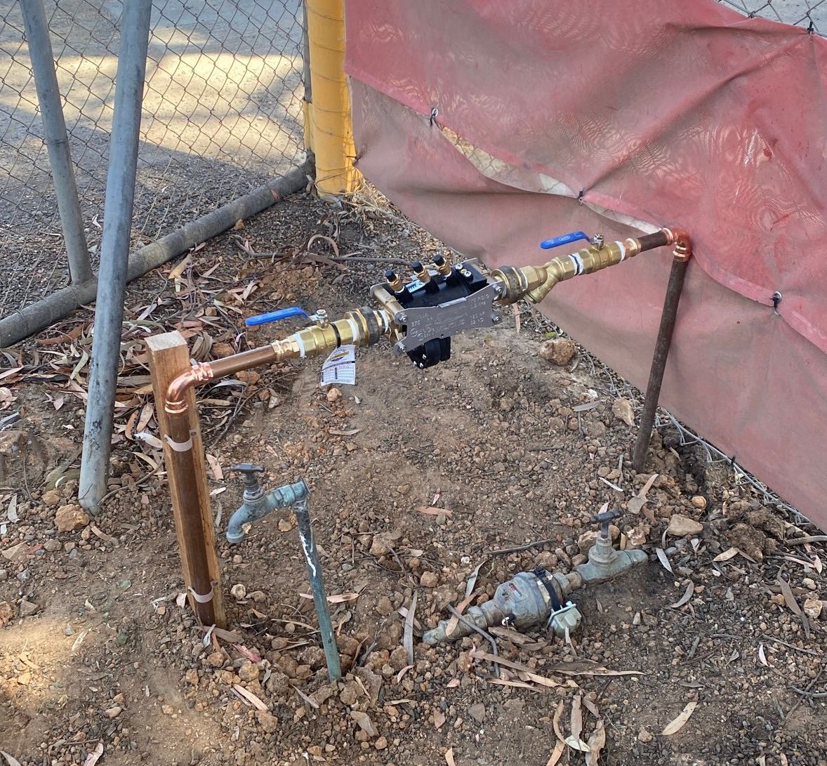Water meter assembly with copper pipes, blue handles, and ground-level spigot, outdoors.