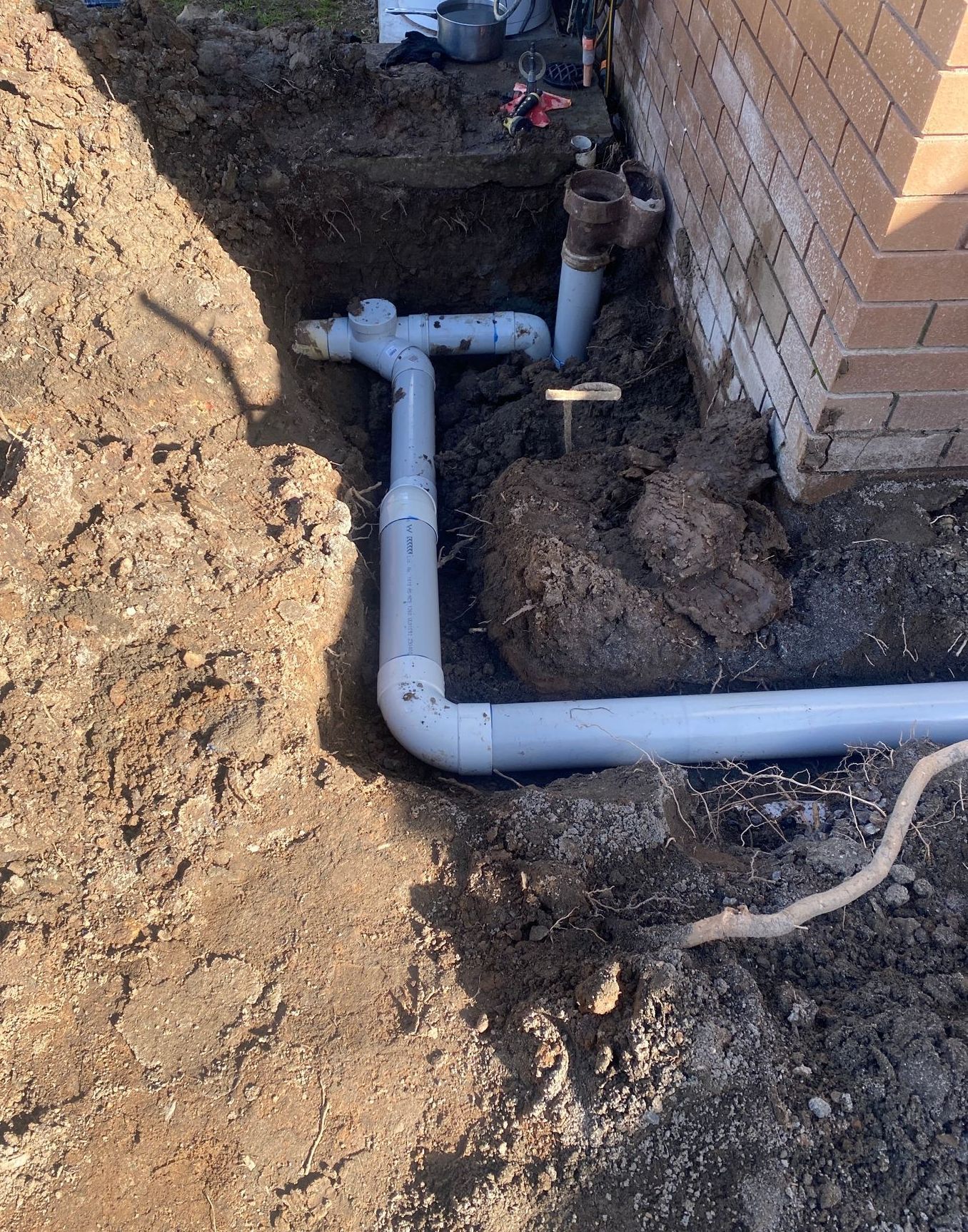 Gray PVC pipes partially buried near a brick wall, likely part of a plumbing or drainage system.