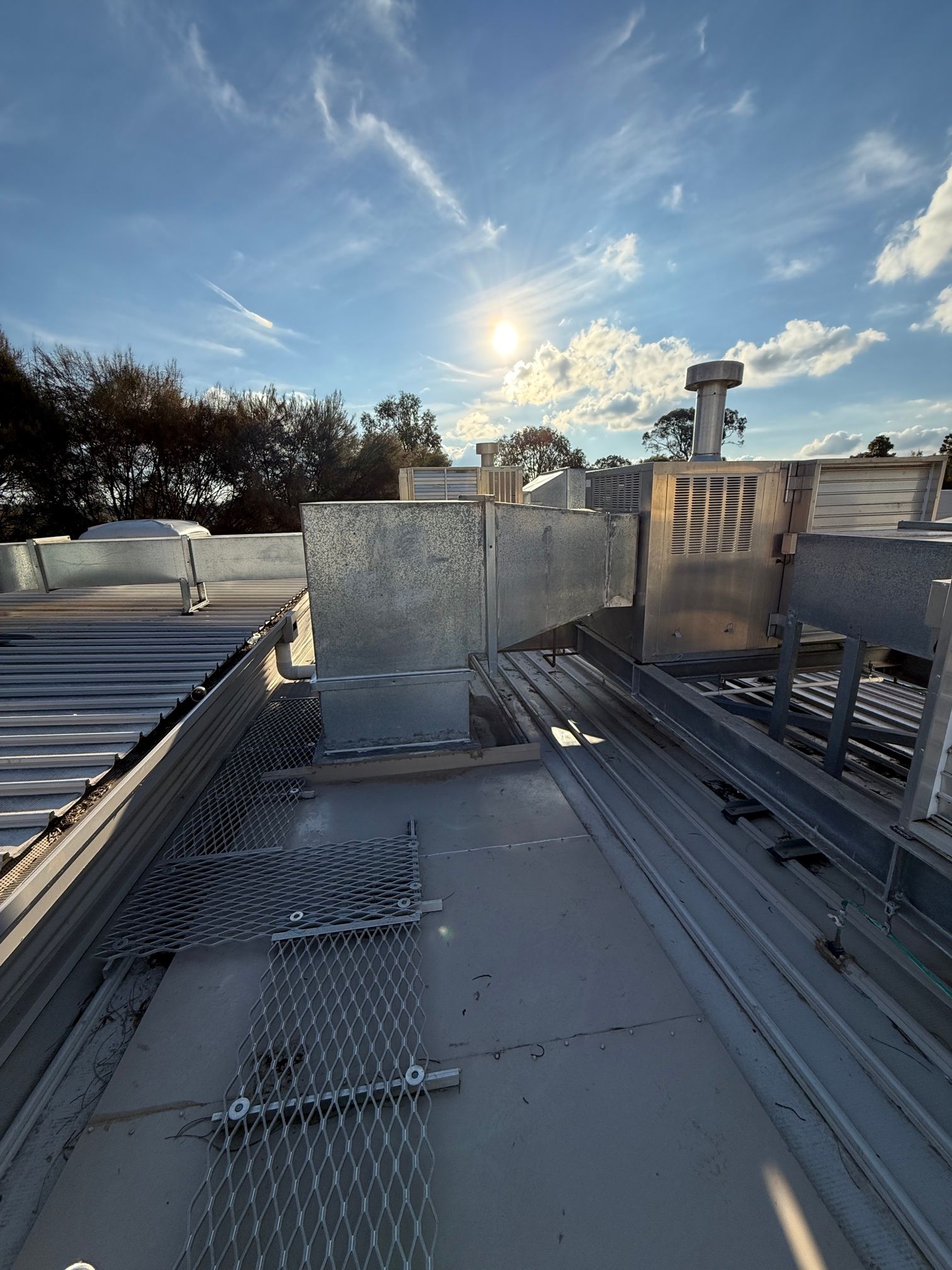 Rooftop with metallic vents, grates, and sunlight, set against a blue sky with visible clouds.