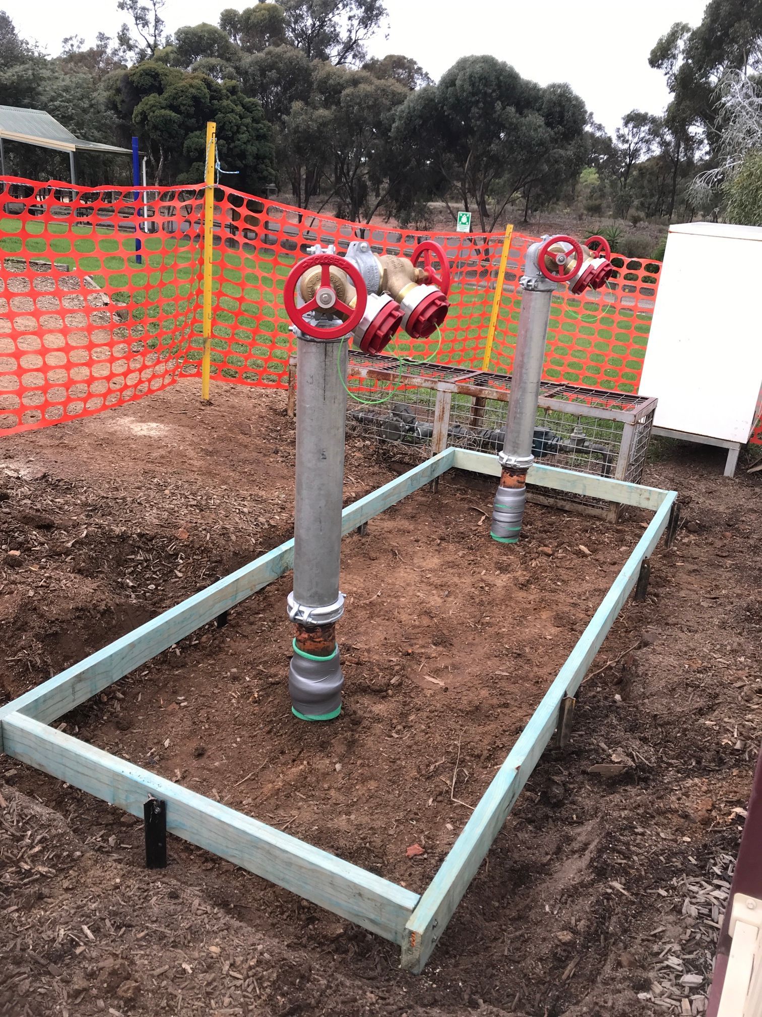 Two vertical pipes with red valves, inside a wooden frame, in a dirt area, with orange fencing.