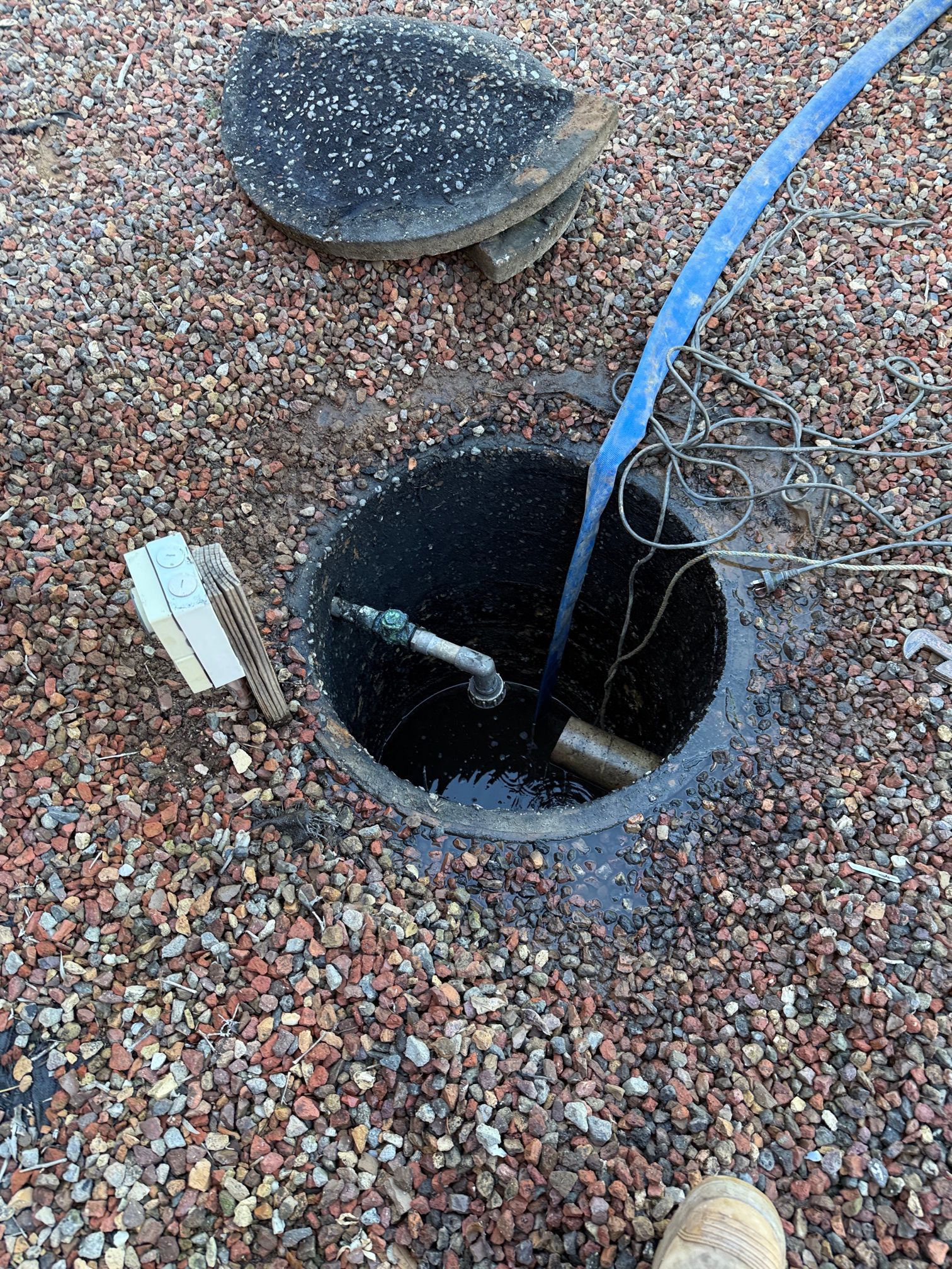 Open manhole with a blue hose, surrounded by gravel. A boot is visible at the bottom.