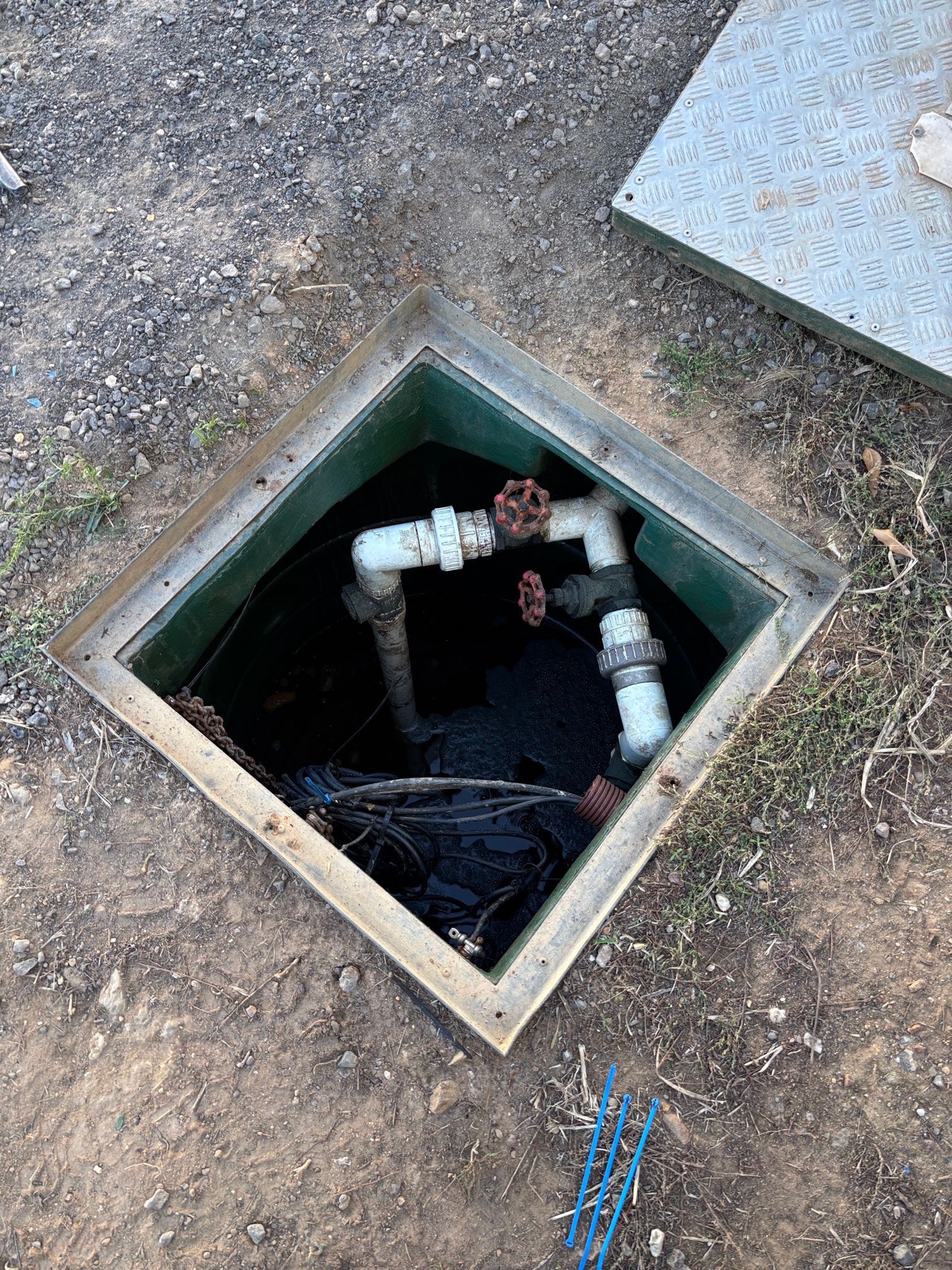 Open utility access box with pipes and valves. Metal cover is partially open, set in the dirt.