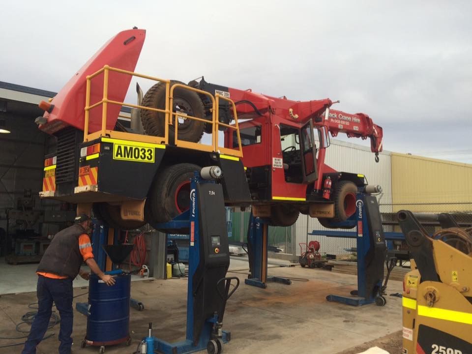 Red and black industrial truck lifted on blue jacks; worker near a blue barrel, outdoors. — RK Diesel Services in Ciccone, NT