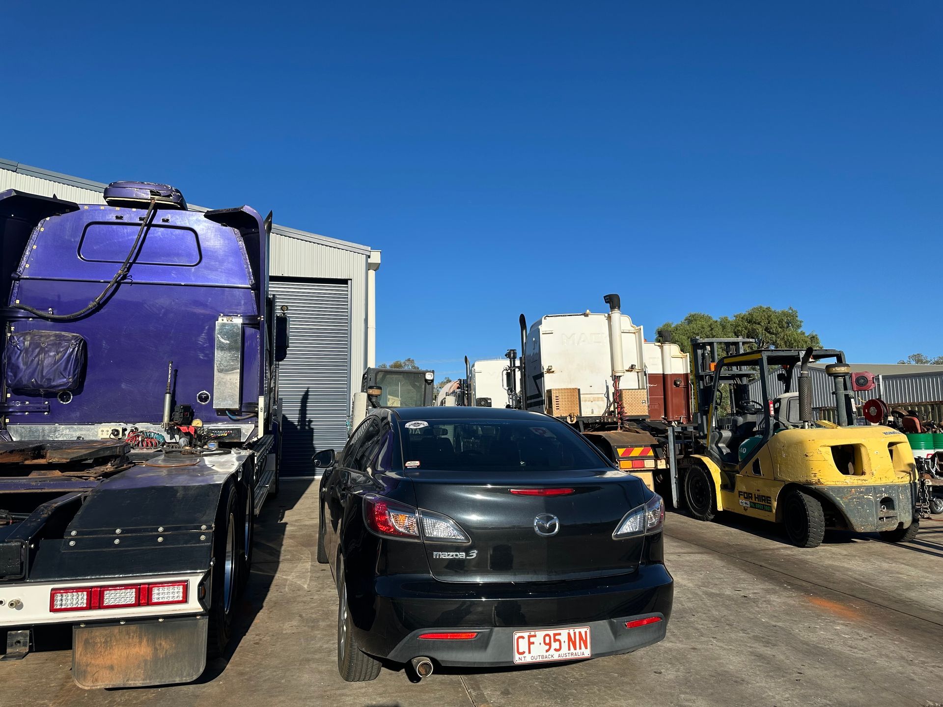 A black Mazda sedan parked near a purple semi-truck and a yellow forklift on a sunny day. — RK Diesel Services in Ciccone, NT