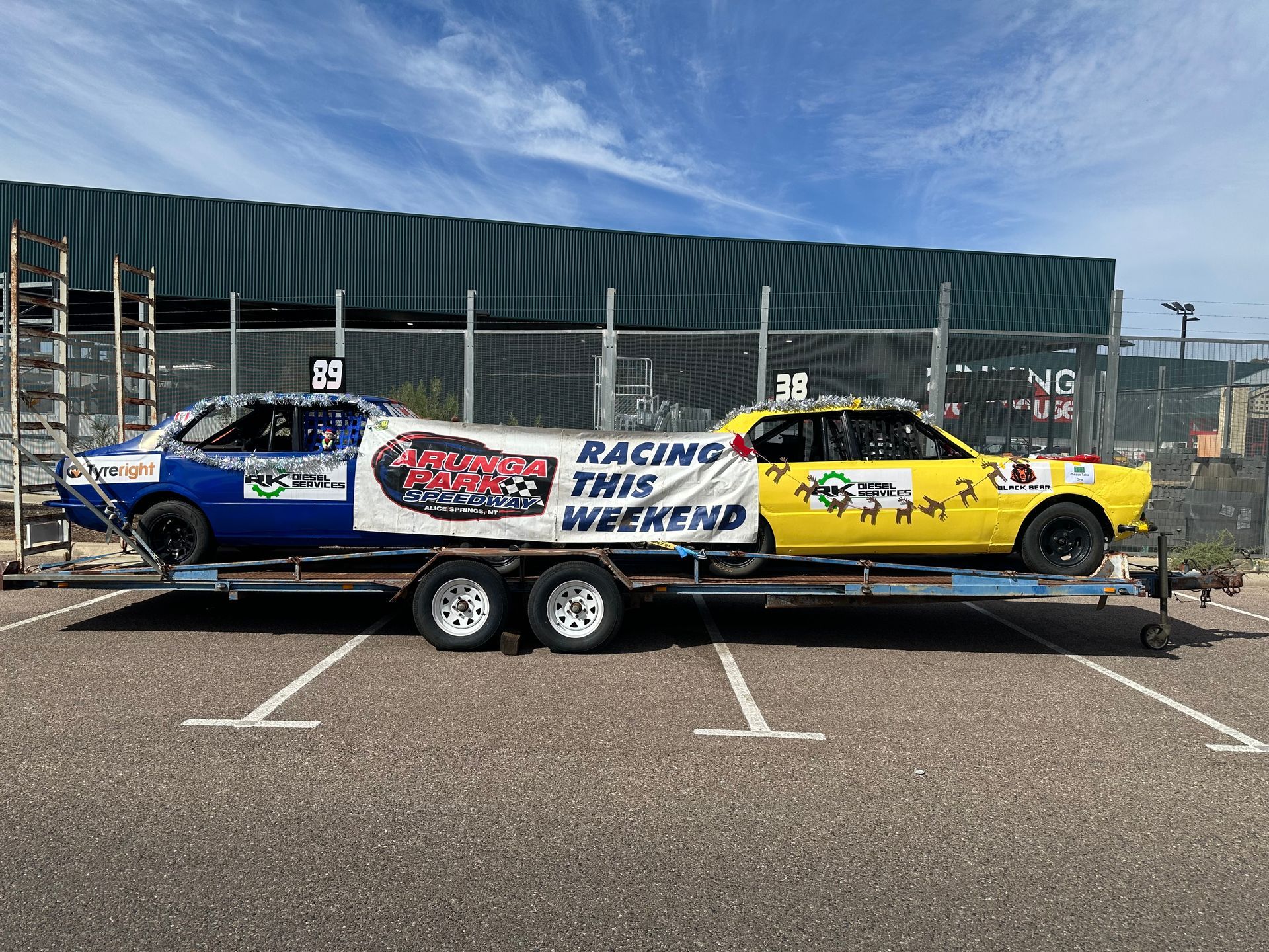 Two race cars on a trailer, one blue and one yellow, with a banner that reads 