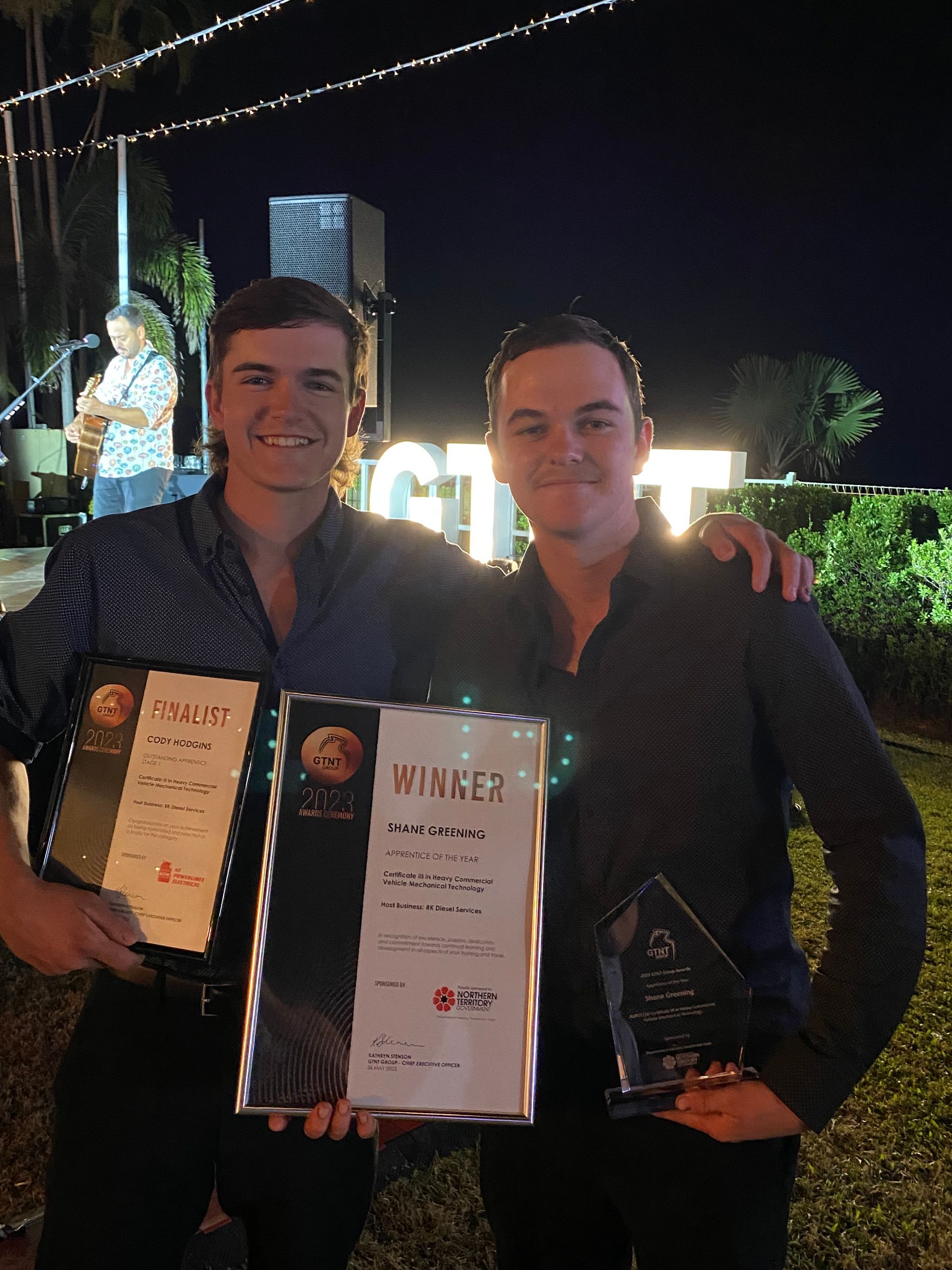 Two men holding awards at an outdoor event, one with a microphone in the background. — RK Diesel Services in Ciccone, NT