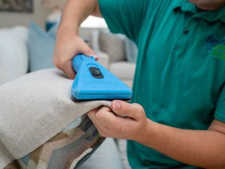 A man in a green shirt is cleaning a couch with a blue tool