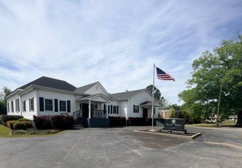 A person waving while sitting atop a black horse-drawn hearse in front of a brick building.