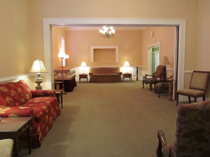 A formal viewing room with a casket, several chairs, and lamps, connected to another room by a wide archway.