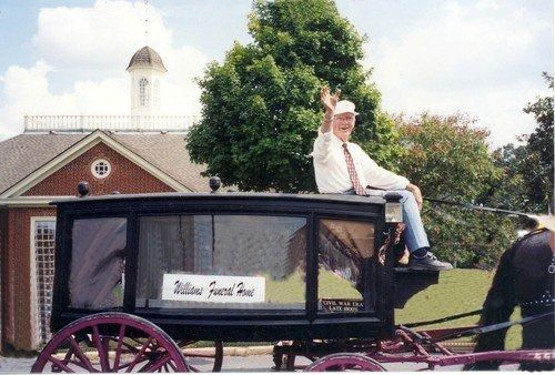 A person waving while sitting atop a black horse-drawn hearse in front of a brick building.