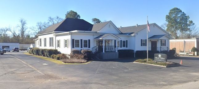 A white, single-story building with a dark roof and black shutters, situated on an asphalt lot under a clear sky.