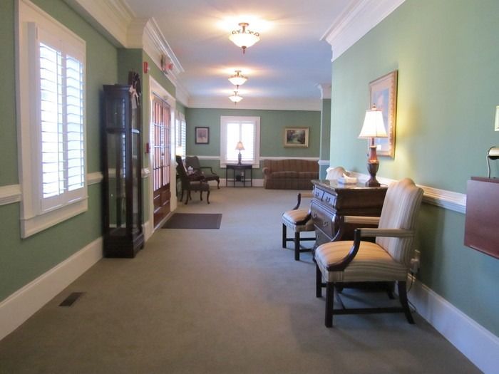 A hallway with sage green walls, light carpeting, three ceiling lights, a tall display case, and chairs along the wall.