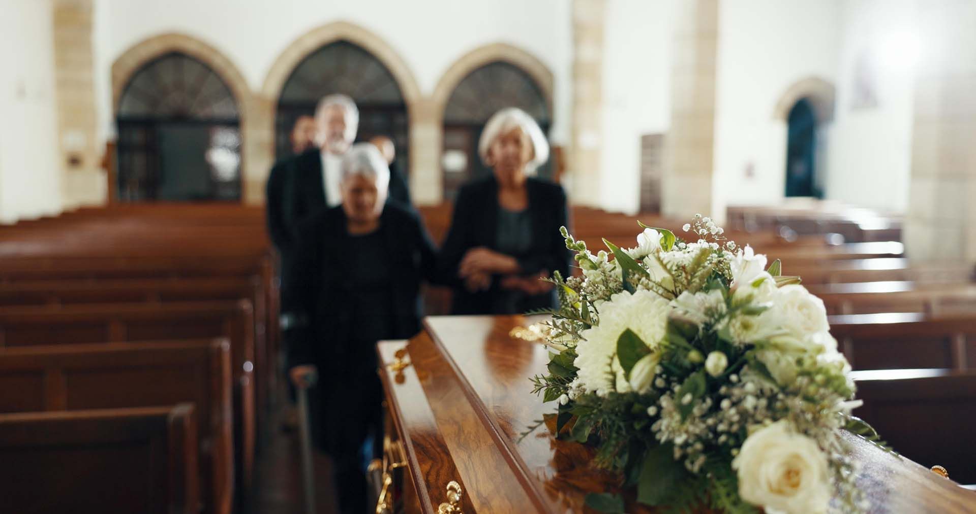 People walking behind a wooden casket adorned with a bouquet of white flowers inside a church.