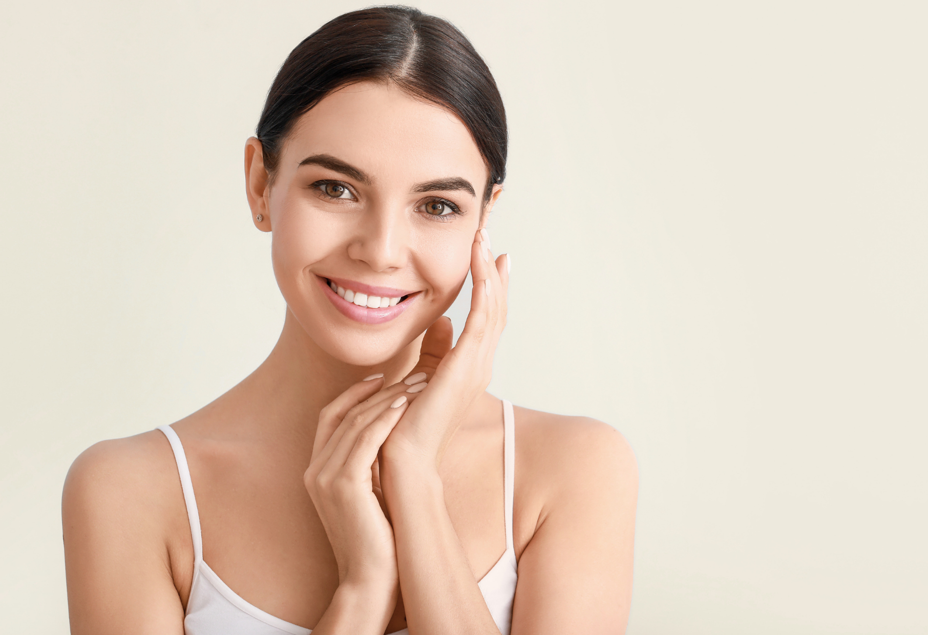 Woman with dark hair in a bun, smiling and touching her face with both hands, white tank top, light background.