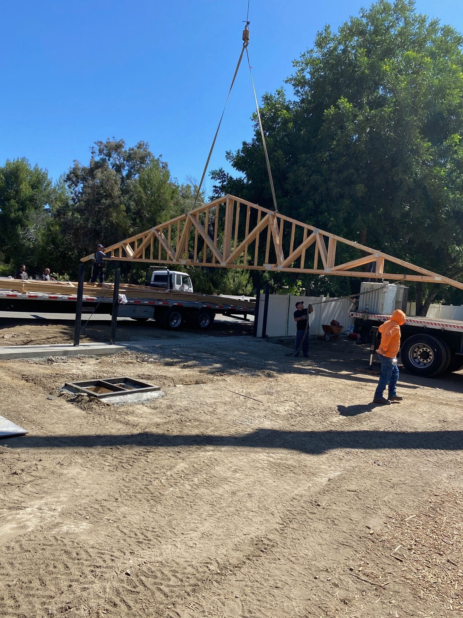 Construction workers lifting a wooden roof truss with a crane at a construction site on a sunny day.