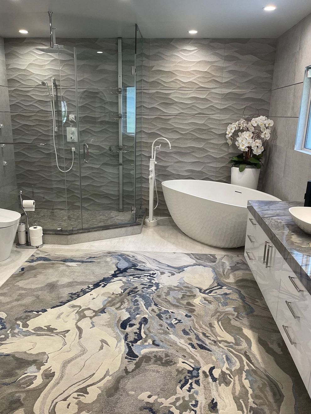 Modern bathroom with gray wave tile, white soaking tub, large patterned rug, and glass shower.